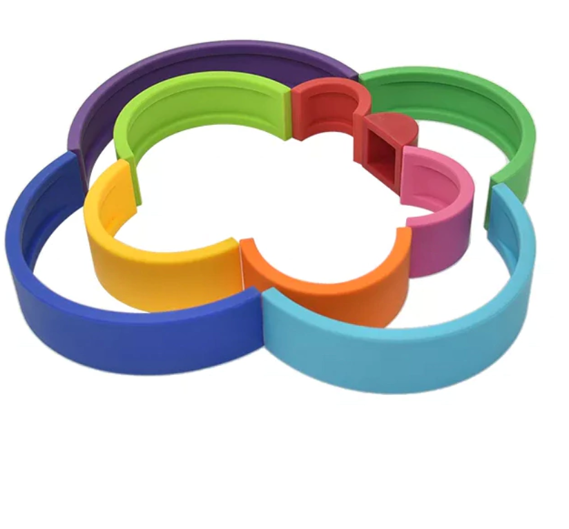 Arco-íris feito de tiras de silicone nas cores vinho, vermelho, rosa, laranja, amarelo, verde limão, verde, azul claro, azul escuro, roxo. Peças desmontáveis para brincar e empilhar e encaixar. Peças colocadas em outro formato para ilustrar ourtos modos de brincar.