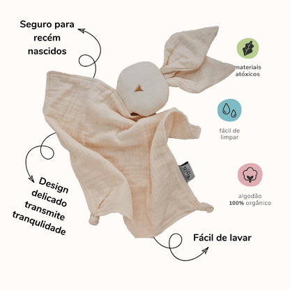 Naninha de algodão em formato de coelho na cor creme com informações sobre produtos. Do lado esquerdo da naninha as seguintes informações: Seguro para recém nascidos, design delicado, transmite tranquilidade. Do lado direito da naninha: materiais átoxicos, fácil de limpar, algodão 100% orgânico, fácil de lavar. 